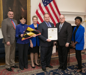 Maryland Governor Larry Hogan, State Superintendent of Schools Dr. Karen B. Salmon with 3 representatives from Urbana High School holding a certificate and a Maryland flag.